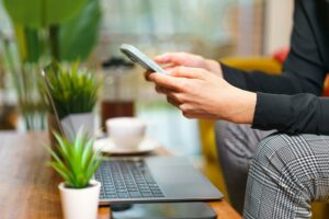 Mains utilisant un smartphone devant un ordinateur portable, illustrant l’externalisation digitale et la productivité